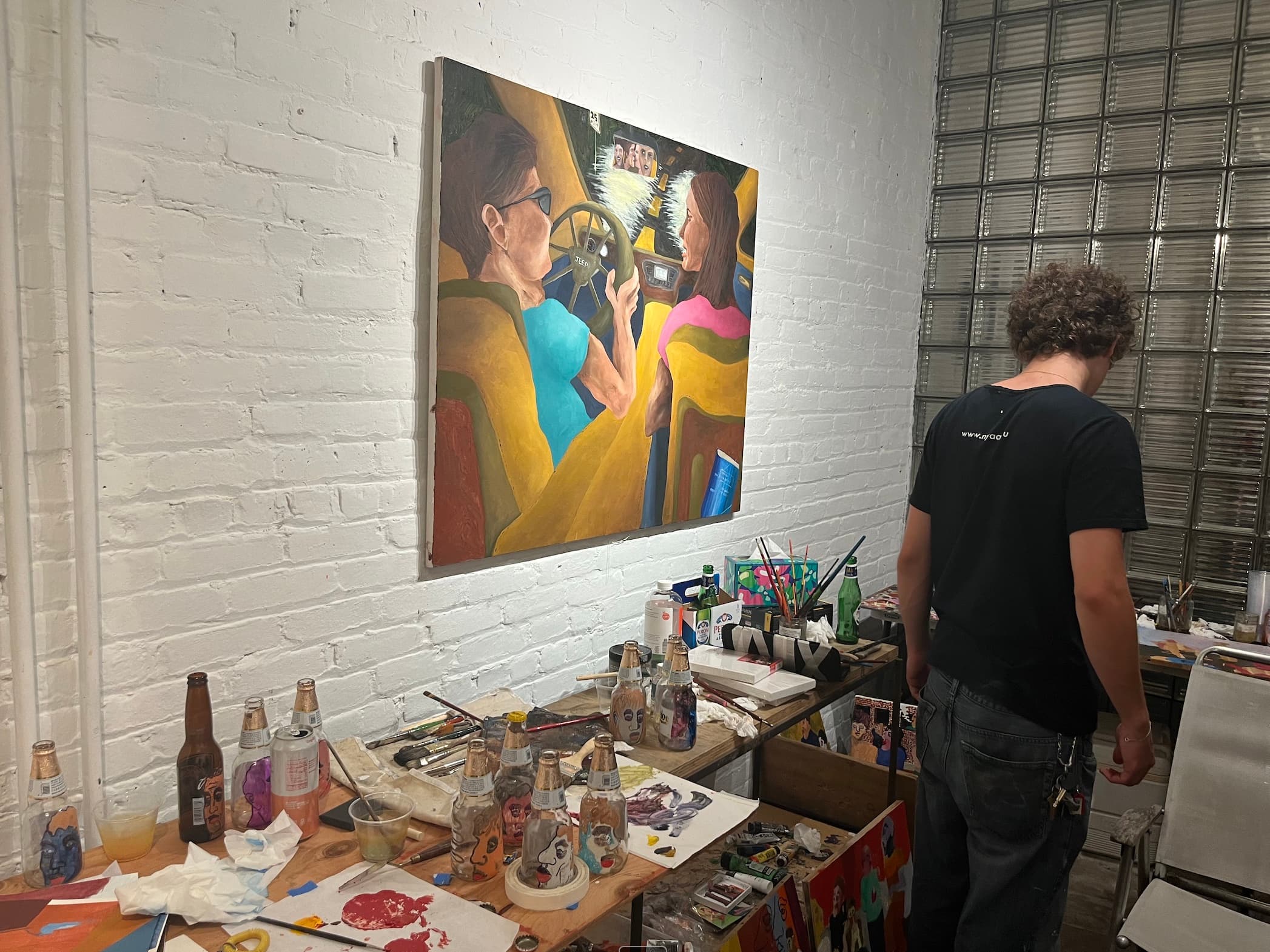 A painting hung on a whitewashed brick wall above a working table of brushes, paint, and hand-painted bottles, with a figure standing over it.
